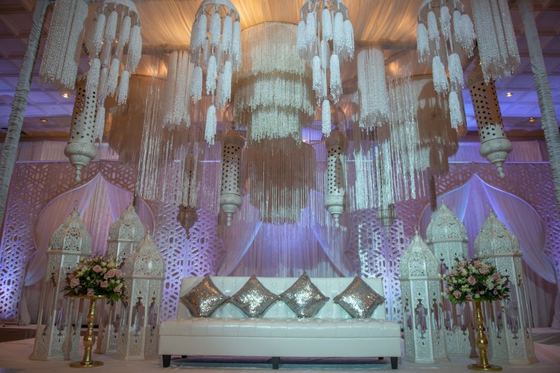 Stage with white Moroccan lanterns, hanging white floral and crystal chandeliers and a tufted white sofa.