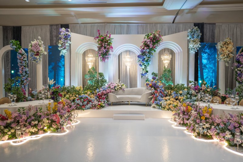 White arch stage with pastel and blue florals, forest-print panels and a curved sofa, edged with lit floral borders.
