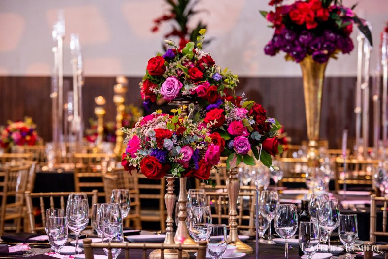 Reception tables with tall gold stands of red, pink and purple roses beside crystal glassware and gold chairs.