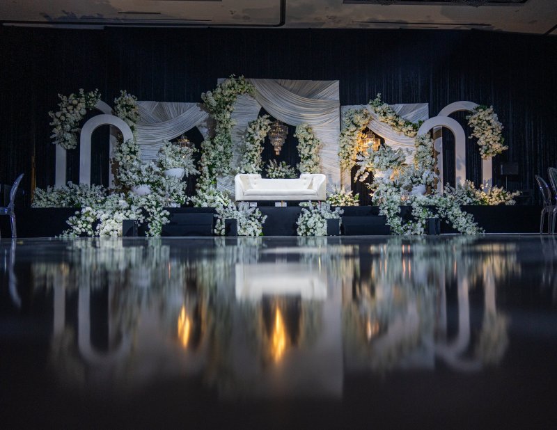 White arch frames with white florals and drapes around a white sofa, reflected in a dark glossy dance floor.