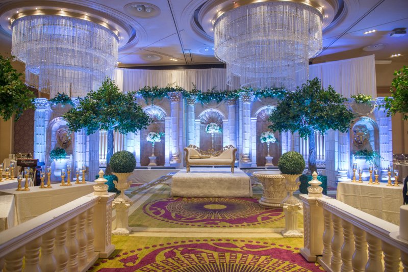 White arched pillar stage with greenery trees and crystal chandeliers, approached by a balustrade aisle over an ornate carpet.