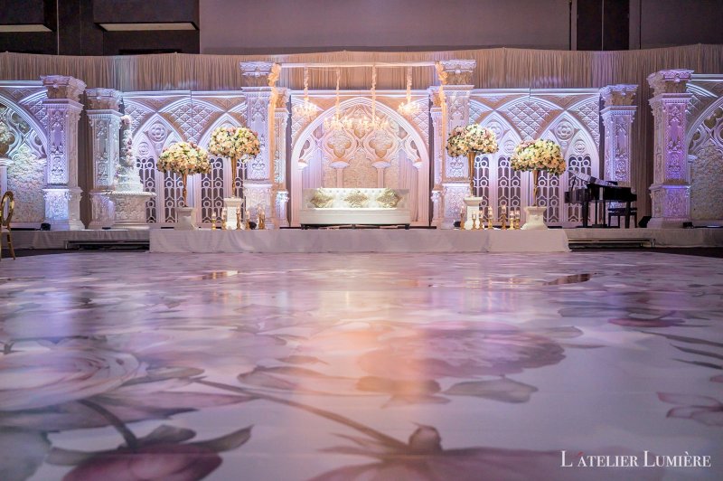White carved panel stage with crystal chandeliers, floral pedestals and a grand piano above a floral-print dance floor.