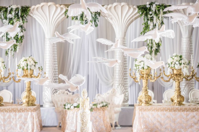 Suspended white dove installation among white palm-style columns with gold candelabras of white roses.