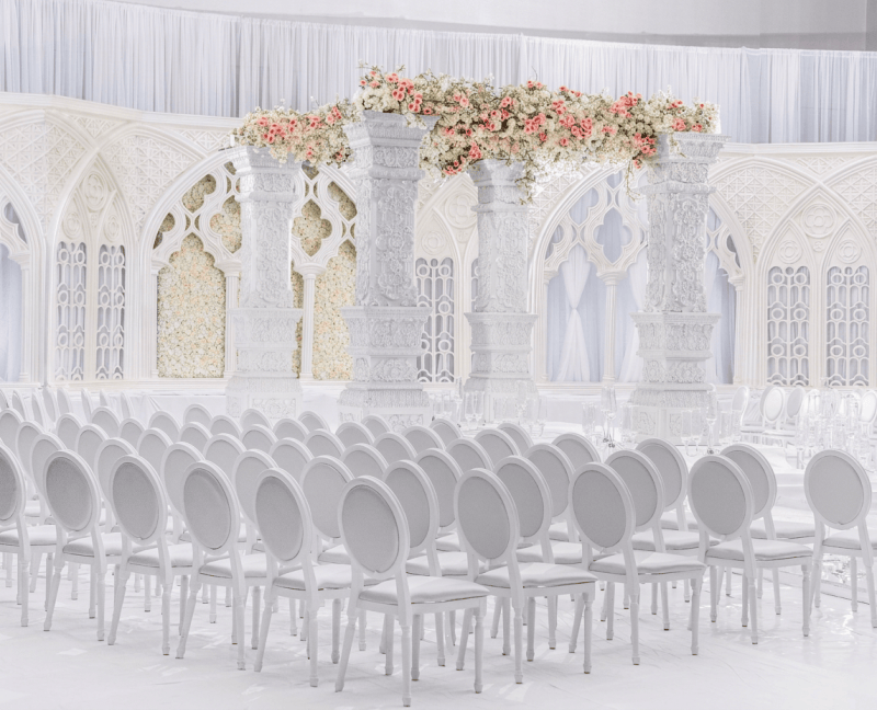 All-white carved pillar mandap with a blush floral canopy, white Gothic-arch panels and rows of white chairs by GPS Decors