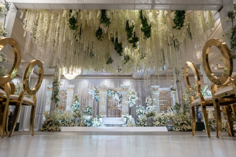 Wedding reception stage with white floral arches under a hanging greenery ceiling, framed by gold chairs, by GPS Decors