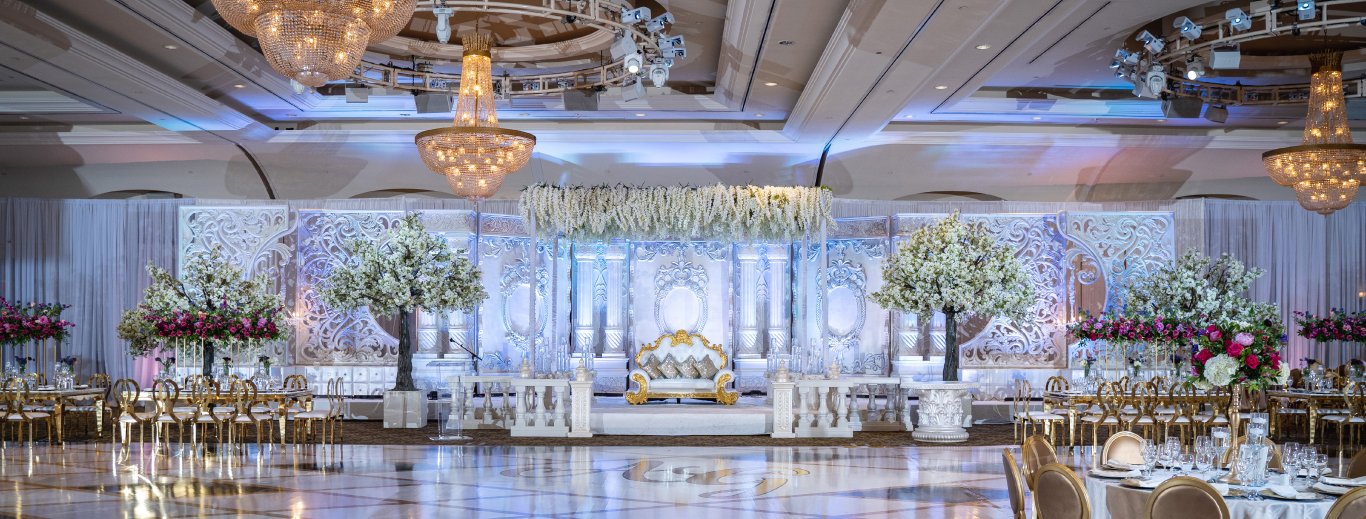 Ballroom reception with a white floral stage, white blossom trees and crystal chandeliers by GPS Decors